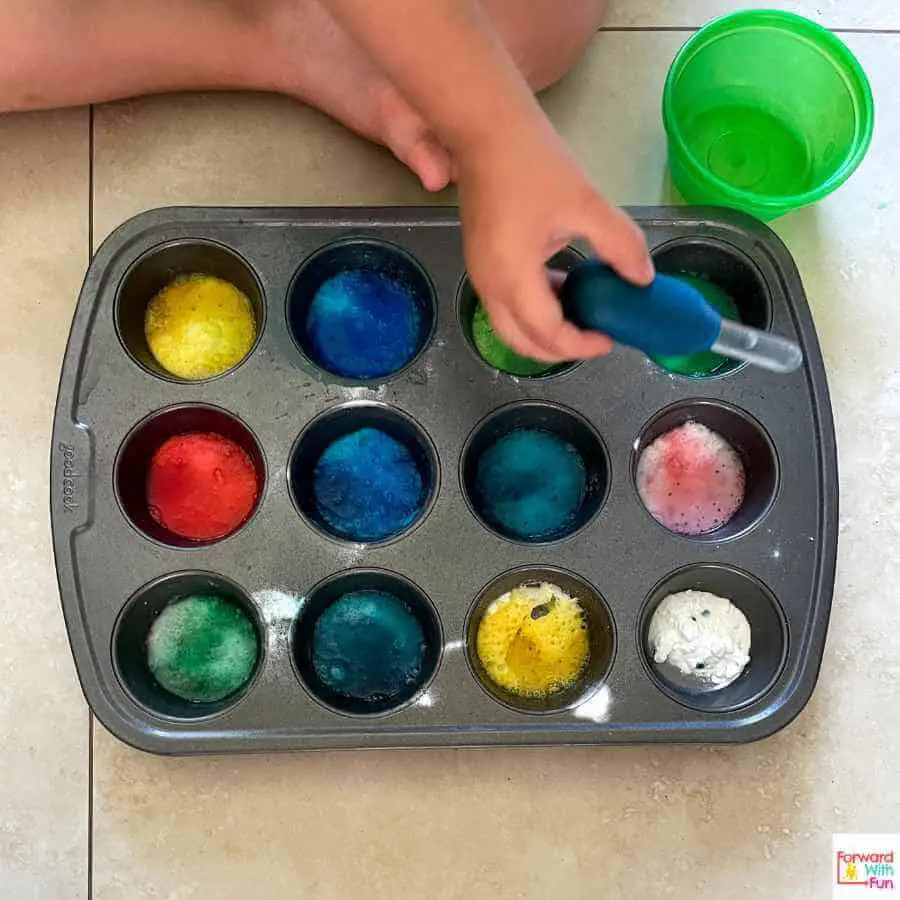 muffin pan with colorful bubbles rising in each cup, last cup has baking soda in it, child's hand holding a pipette with white vinegar in it